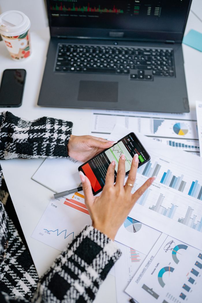 Person analyzing business charts with smartphone and laptop on desk.